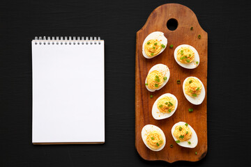 Homemade Deviled Eggs with Chives, blank notepad on a rustic wooden board on a black surface, top view.