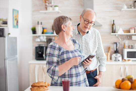 Senior woman holding smartphone and sharing the recipe with husband. Caucasian senior wife and husband in kitchen during breakfast using smartphone technology and internet conection. Leisure retired - Powered by Adobe