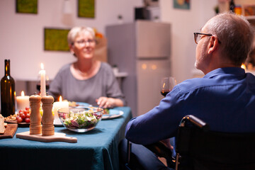 Handicapped man holding whine glass sitting in wheelchair during festive dinner. Happy cheerful senior elderly couple dining together in the cozy kitchen, enjoying the meal, celebrating their