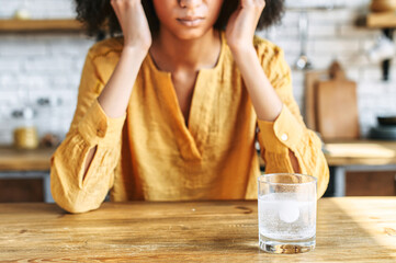 A glass of water with effervescent tablet on the table. Drug, remedy from pain. A woman holds temples with hands on the background, a face is not visible