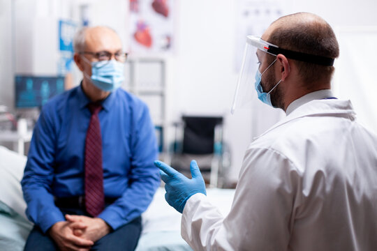 Senior Man With Face Mask Agasint Covid Dicussing Treatment With Doctor In Clinic Cabinet. Global Health Crisis, Medical System During Pandemic, Sick Elderly Patient In Private Hospital.