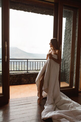 Young woman enjoy morning in hotell room in mountains. She just wake up, covered in white blanket stand on the balcony and enjoy nature view