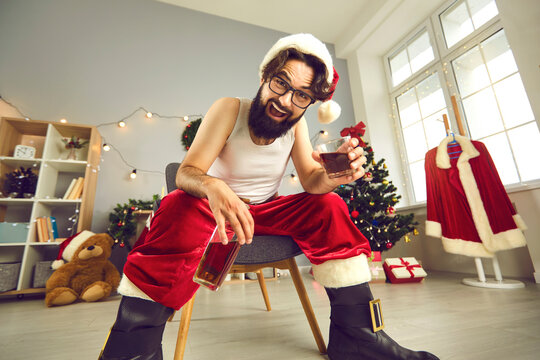 Happy man in undershirt and Santa costume drinking whiskey at Christmas party at home
