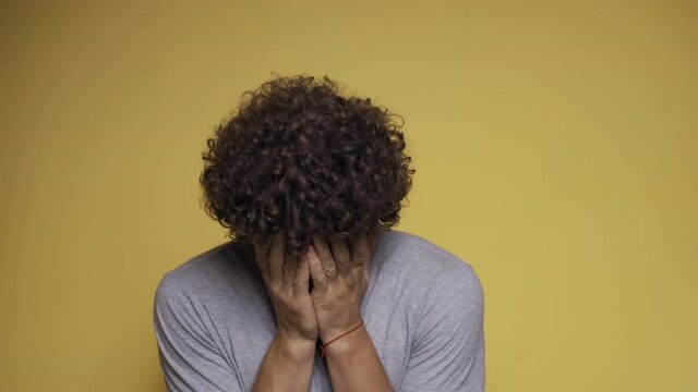 Curly-haired guy sobs with his hands covering his face. Man cries in the studio against an isolated background. Convulsive weeping. High quality 4k footage.