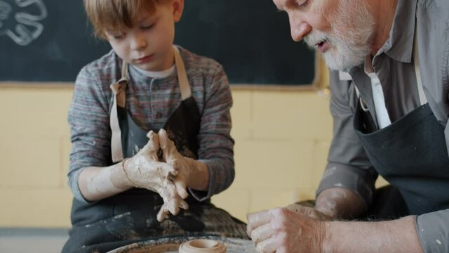 Professional potter is teaching little student to work with throwing-wheel during pottery class indoors in modern studio. Creative hobby and arts concept.