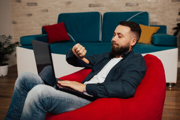 Young bearded man working using laptop sitting on red armchair with angry face, negative sign showing dislike with thumbs down.