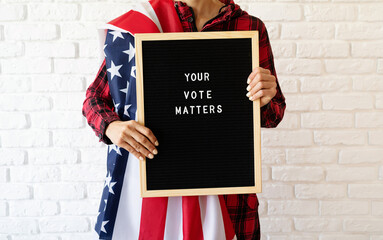 woman with american flag holding letter board with text Your Vote Matters on white brick background