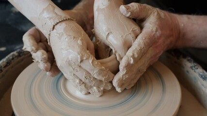 Close-up of adult man's hands helping kid during craftswork on throwing wheel shaping clay in ceramic vase. People and traditional arts concept.