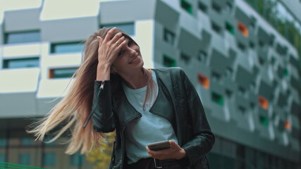 Good looking girl with Long Brown Hair Looking at Her phone Typing and Smiling. Enormous industrial Building at the Background. Green Bushes and Trees. Smart clothes. Natural makeup.