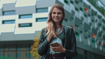 Good looking girl with Long Brown Hair Looking at Her phone Typing and Smiling. Enormous industrial Building at the Background. Green Bushes and Trees. Smart clothes. Natural makeup.