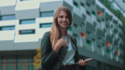 Good looking girl with Long Brown Hair Looking at Her phone Typing and Smiling. Enormous industrial Building at the Background. Green Bushes and Trees. Smart clothes. Natural makeup.