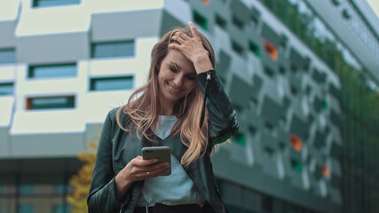 Good looking girl with Long Brown Hair Looking at Her phone Typing and Smiling. Enormous industrial Building at the Background. Green Bushes and Trees. Smart clothes. Natural makeup.