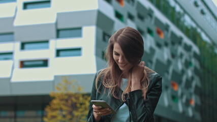 Good looking girl with Long Brown Hair Looking at Her phone Typing and Smiling. Enormous industrial Building at the Background. Green Bushes and Trees. Smart clothes. Natural makeup.