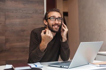 A positive man with a beard is talking on a cell phone and gesturing. Guy with glasses works at home