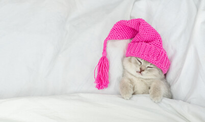 Tiny kitten wearing warm hat sleeps under white blanket on a bed at home. Top down view