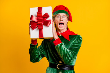 Photo of young funky man open mouth hold giftbox red bow wear elf velvet green costume cap glasses...