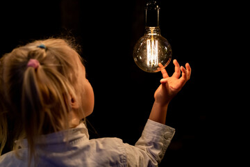 Cute adorable caucasian blond girl portrait smiling and holding in hand one of hanged edison light bulb at forest outdoor. Right solution choice concept