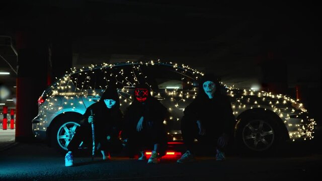People with luminous purge masks are holding a baseball bat and katana. Killer at night in the parking lot against the backdrop of a car with a garland.