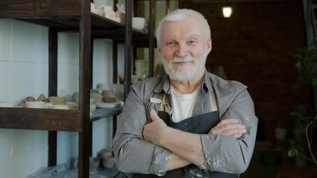 Portrait of cheerful senior ceramist standing in workshop wearing apron and smiling looking at camera. Traditional crafts and creative people concept.