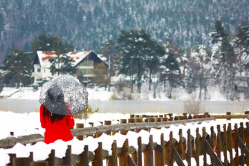 A view of alone and sad girl in red coat and black hair with umbrella walking on a wooden road  against the forest by the lake.