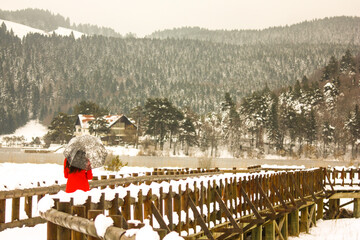 A view of alone and sad girl in red coat and black hair with umbrella walking on a wooden road  against the forest by the lake.