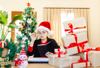 Obraz na płótnie Canvas Woman in hat santa office preparing for Christmas,ckecking list, many presents and christmas tree in background, laptop on table, working New Year desk at home