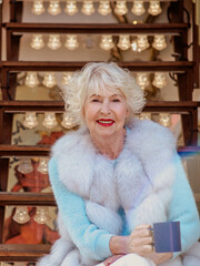 senior stylish woman in fur coat and with grey hair sitting on carousel drinking tea (coffee) and...