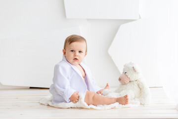 a small fat baby six months old in a white jacket and diapers sits on a light background and plays with a Teddy bear, space for text