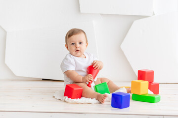 cute little baby six months old in a white t shirt and diapers playing at home on a Mat in a bright room with bright colored cubes