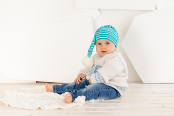 a small happy child six months old in a knitted warm jacket and a cap is sitting at home on a rug in a bright room