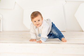 a small happy child six months old in a warm knitted jacket and blue jeans crawls at home in a...