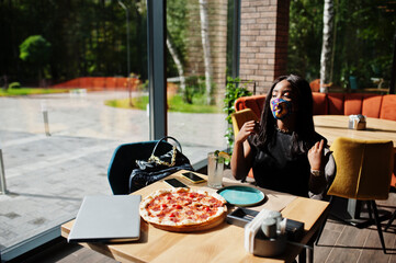 Fashionable african american woman, wear hand made face mask sitting at restaurant while eat pizza....