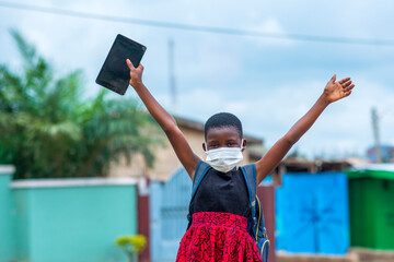 Portrait of an African child student wearing face mask and holding digital tablet with arms raised up - concept on education in covid-19 pandemic