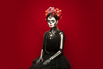 Scary. Young girl like Santa Muerte Saint death or Sugar skull with bright make-up. Portrait isolated on red studio background with copyspace. Celebrating Halloween or Day of the dead.