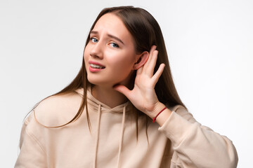 Young pretty brunette woman keeps palm of a hand close to ear as if listening to rumors isolated...