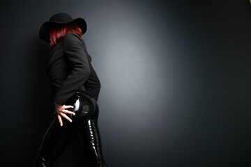 Fototapeta na wymiar Beautiful fashion woman with perfect body, back view. Portrait of beautiful girl wearing black blazer, latex tight pants and hat posing in studio over deep gray background