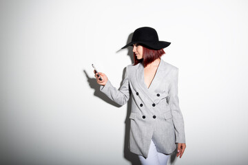 Young stylish woman using smartphone, indoor portrait over white. Fashion modern girl wearing grey jaket and black hat texting on her mobile phone