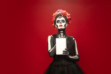 Book. Young girl like Santa Muerte Saint death or Sugar skull with bright make-up. Portrait isolated on red studio background. Celebrating Halloween or Day of the dead. Copyspace on cover.