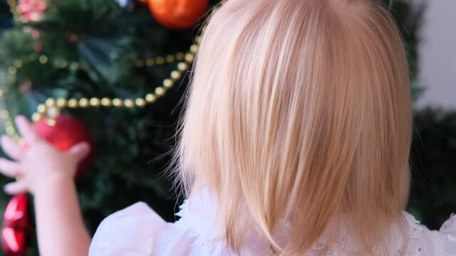 baby, a little blonde girl in a white dress, plays with New Year's decorations, examines Christmas balls, concept of waiting for gifts, festive mood