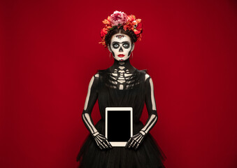Holding tablet. Young girl like Santa Muerte Saint death or Sugar skull with bright make-up. Portrait isolated on red studio background. Celebrating Halloween or Day of the dead. Copyspace on screen.
