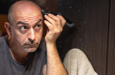 A middle-aged Caucasian man cuts his hair in the bathroom. While placing the clipper on your head check with your eyes in the mirror.