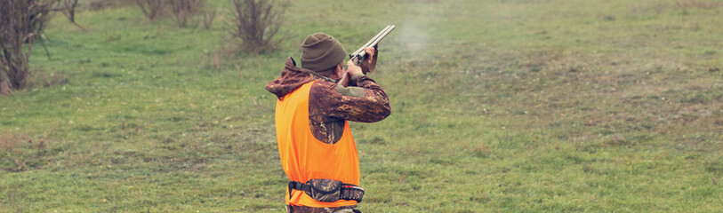 Fototapeta premium A man with a gun in his hands and an orange vest on a pheasant hunt in a wooded area in cloudy weather. Hunter with dogs in search of game.
