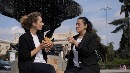 Lunch break outdoors. Two female office workers eating fast food near the fountain 