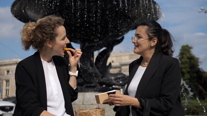 Two female co-workers enjoying their lunch break outdoors with take away meal their share next to the fountain. Maintaining healthy workplace