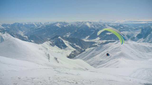 Beautiful epic landscape of high altitude mountains, bright coloured paraglider parachute flies down. Amazing scene of extreme sports for winter activity at ski resort