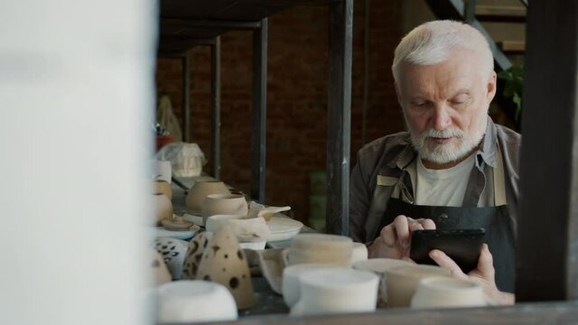 Slow motion of elderly man potter using tablet during inventory in workshop counting ceramics and touching screen of modern device. Technology and business concept.