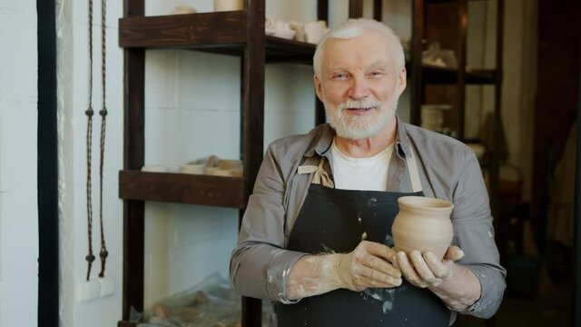 Portrait of senior potter in muddy apron standing in workshop holding pot smiling and looking at camera. Traditional crafts and professionals concept.