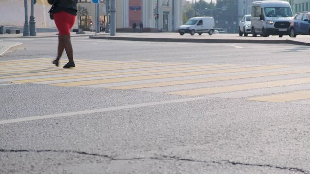 Close-up feet of people walking along the crosswalk on a sunny day. Cars are moving in the background out of focus.