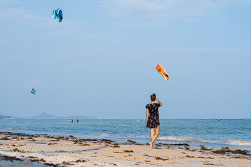 Young woman in dress stands on the sandy beach at sunset and looks at the kite-surfers in the sea 