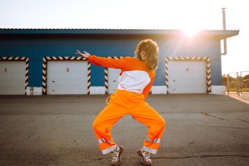 Young sporty woman dancer dancing in the street at sunset. African American woman in an orange suit showing some moves. Sport, dancing and urban culture concept. - obrazy, fototapety, plakaty